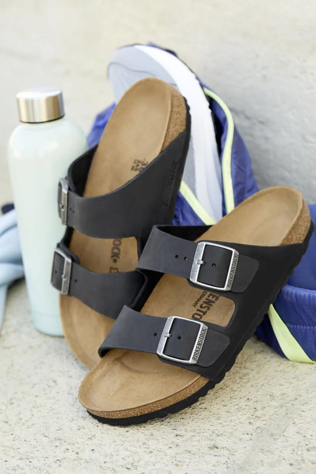Black Birkenstock Arizona sandals displayed beside a backpack and water bottle.